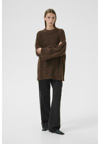 Oversized bruine gebreide trui met lange mouwen, ronde hals en zichtbare textuur; gecombineerd met zwarte wijde broek en puntige schoenen.