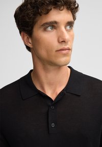 Polo nero realizzato in tessuto texturizzato, con colletto classico e pattina a tre bottoni. La vestibilità è slim e moderna.