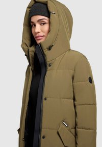Padded olive green jacket with a large hood, black zipper, and snap button accents. Features a side pocket and quilted texture.