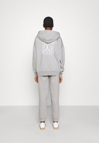Personne aux cheveux tressés portant un sweat à capuche gris et un pantalon de survêtement assorti, baskets blanches, debout face à un mur blanc, les mains détendues le long du corps.