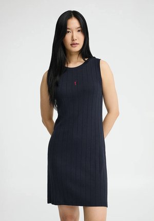 Junge Frau mit langen schwarzen Haaren, die ein ärmelloses, geripptes, marineblaues knielanges Kleid mit kleinem rotem Logo auf der Brust trägt.