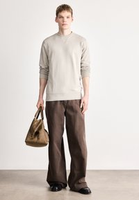 Φωτεινό μπεζ φούτερ με ριμπ μανσέτες, συνδυασμένο με φαρδιά καφέ παντελόνια. Το μοντέλο κρατάει μια καφέ τσάντα με στρογγυλεμένο σχήμα και κοντά χερούλια.