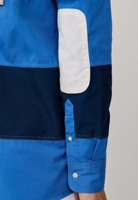 Overhemd met knoopsluiting, uitgevoerd in gradiënt blauw panels, een witte elleboogpatch en zachte katoen. Inclusief knoopsluitingen op de mouwen.