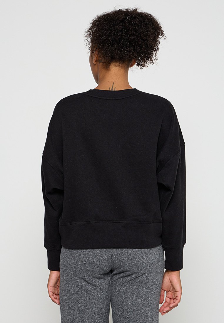 Frau mit lockigem Haar in einem schwarzen Langarm-Sweatshirt und grauen Hosen, die vor einem schlichten weißen Hintergrund steht und die Hände entspannt an den Seiten hält.