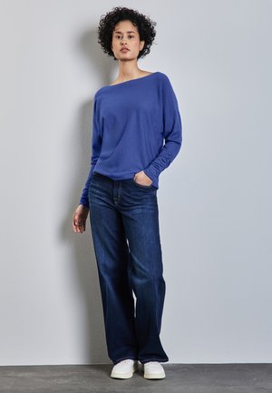Blauwe langemouwen top met een bootnek, gecombineerd met donkerblauwe wijde jeans en witte sneakers, tegen een neutrale achtergrond.