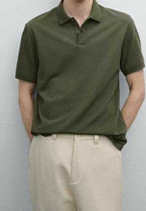 Olivgrünes Polohemd mit Kragen und kurzen Ärmeln, aus strukturiertem Stoff; getragen mit hellbeigen Hosen, die einen Knopfdetails aufweisen.