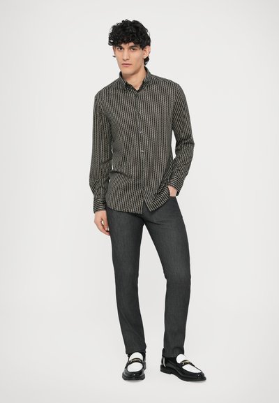 Juoda marškiniai su baltomis vertikaliomis juostelėmis, ilgomis rankovėmis ir klasikiniu apykakliu, derinami su tamsiais džinsais ir blizgančiais juodais batais.