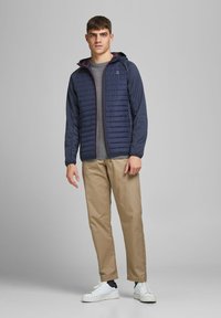 Navy quilted jacket with red and blue accents, gray sweater, khaki trousers, and white sneakers. Model stands in a neutral background.
