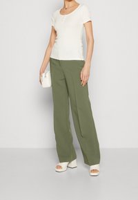 Valkoinen lyhythihainen pusero, jossa on syvä pyöreä pääntie ja kolme nappia, yhdistetty korkeavyötäröisiin oliivinvihreisiin leveälahkeisiin housuihin ja kermaisiin muhkeisiin sandaaleihin.