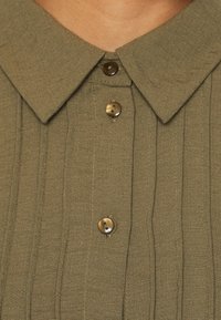 Nahaufnahme eines olivgrünen, gerippten Stoffhemds mit spitzen Kragen und drei sichtbaren, runden braunen Knöpfen.