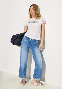 Weißes T-Shirt mit kurzen Ärmeln und dem Text "NEW LINES", hellblaue weit geschnittene Jeans und silberne Sandalen. Glattes Material und lässige Passform.