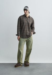 Brūns rūtainas pogas uz augšu krekls, gaiši zaļas platām kājām bikses un brūnas rakstainas čības. Modelis valkā pelēku cepuri ar grafisku dizainu.