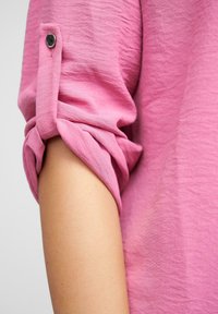 Manica di camicia rosa chiaro con polsini arrotolati fissati da un bottone scuro. Tessuto morbido e testurizzato con pieghe sottili. Pelle liscia visibile sotto.