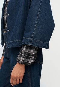 Άτομο που φοράει σκούρο μπλε τζιν μπουφάν και παντελόνι πάνω από ένα ασπρόμαυρο πουκάμισο καρό, με τα μανίκια στρωμένα και να ακουμπούν κοντά στο πλευρό του.