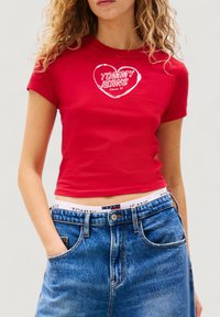 Maglietta rossa corta con un logo bianco a forma di cuore con la scritta "TOMMY JEANS Since 85", abbinata a pantaloncini di jeans blu a vita alta.