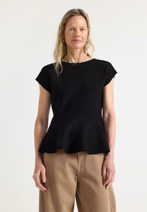 Donna con capelli biondi lunghi che indossa una maglia nera a maniche corte con orlo a balze e pantaloni beige a gamba larga, su sfondo semplice.