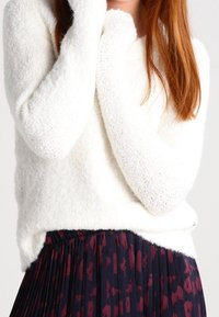 Personne aux longs cheveux roux portant un pull blanc duveteux et une jupe plissée marine avec des motifs rouge foncé, les bras croisés près du visage.