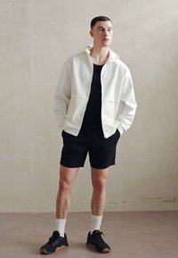 Un sweat à zip blanc avec deux poches avant, associé à un débardeur noir et un short noir. Des baskets noires avec une semelle en caoutchouc, des chaussettes blanches montantes.