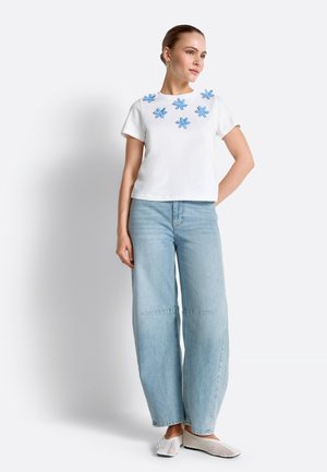 Kobieta w białej bluzce z krótkim rękawem ozdobionej niebieskimi aplikacjami kwiatowymi, szerokich jasnoniebieskich dżinsach i białych butach wsuwanych, stojąca na jednolitym tle.