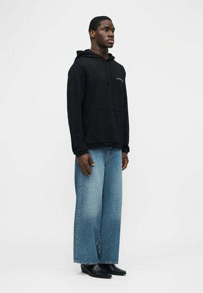 Άνδρας που στέκεται φορώντας μαύρο φούτερ με κουκούλα και μπροστινή τσέπη, φαρδιά ξεθωριασμένα μπλε τζιν και μαύρα μυτερά παπούτσια μπροστά σε απλό φόντο.