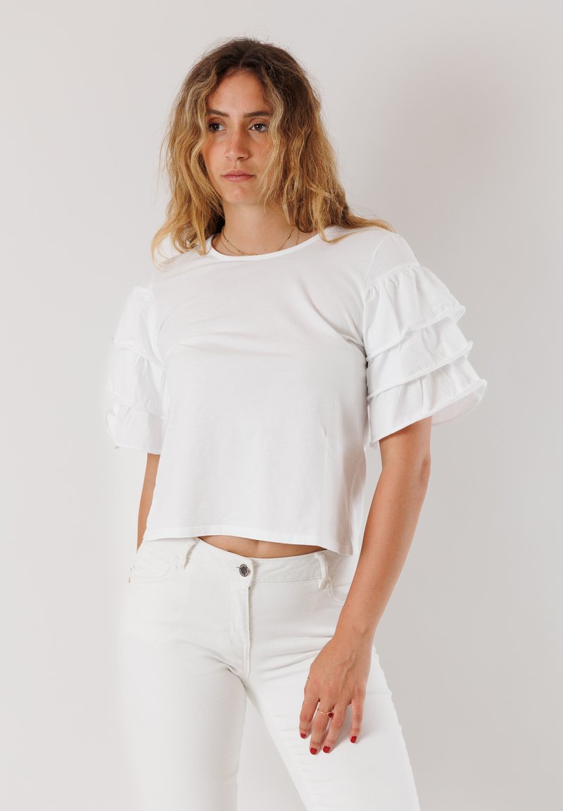 T-shirt bianco cropped con maniche corte arricciate. Realizzato in un tessuto morbido, presenta un collo rotondo e una vestibilità ampia. Abbinato a jeans bianchi.