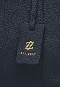 Etichetta in pelle nera testurizzata con logo intrecciato dorato "ZZ" e testo "EST. 2007" attaccata a una superficie in pelle nera cucita.