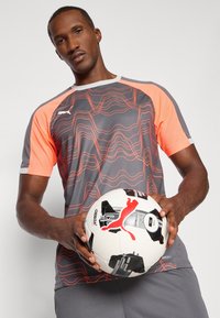 Sivý športový dres s jasne oranžovými sieťovanými rukávmi a vzorom vlnitej línie; držiaci futbalový loptu má čierne akcenty a logo.