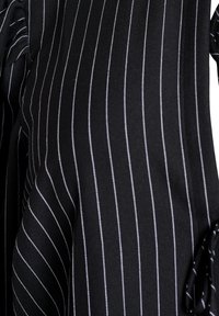 Črn material z belimi navpičnimi črtami, ki ima teksturiran tkan. Vključuje trakove na straneh za prilagodljivost.