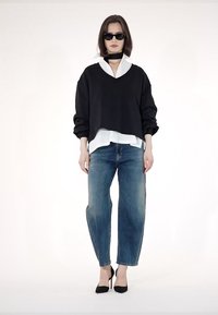 Crni oversized pulover složen preko bijele košulje, plavih traperica i crnih štikli; model nosi tamne sunčane naočale i choker.