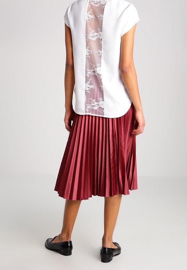 Haut blanc à manches courtes avec un dos en dentelle transparente. Jupe plissée bordeaux fluide. Chaussures plates noires complètent la tenue.