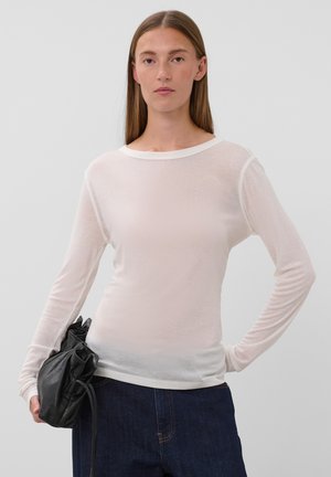 Femme aux cheveux bruns et raides portant un haut blanc transparent à manches longues et un jean bleu foncé, tenant un sac en cuir noir à ses côtés.