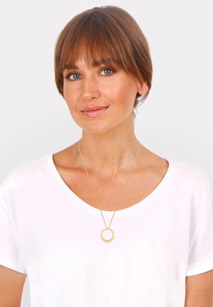 Goldener runder Anhänger-Halskette an einer dünnen Kette, getragen über einem schlichten weißen T-Shirt. Einfaches Design, glatte Textur, minimalistischer Stil.