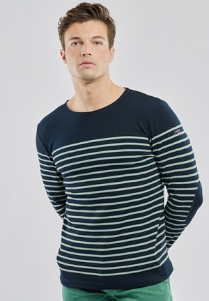 Jeune homme aux cheveux courts portant un t-shirt à manches longues rayé horizontalement bleu marine et vert clair et un pantalon vert, debout devant un fond uni.