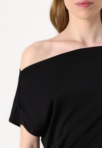 Juodas off-shoulder marškinėliai, pagaminti iš lygaus, elastingo audinio. Pasižymi laisvu siluetu, kabinamu dizainu ir minimaliu siūlių skaičiumi, siekiant švaraus įvaizdžio.