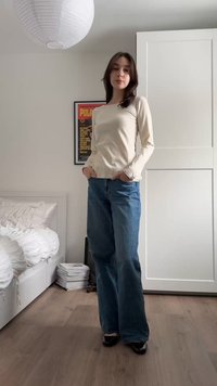 Tânără cu bluză bej cu mâneci lungi și blugi albaștri, stând în dormitor cu mâinile în buzunare, pat alb și un dulap în fundal.