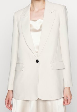 Vrouw die een crèmekleurige blazer met één knoop draagt over een satijnen crèmekleurige top en bijpassende rok, staand tegen een effen achtergrond.