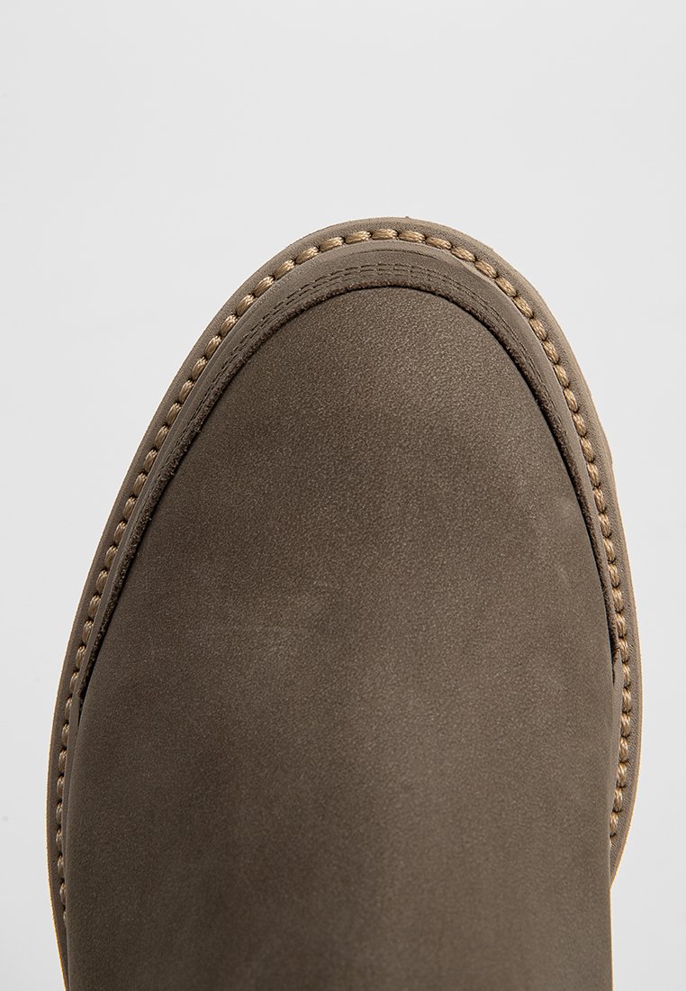 Braune Wildlederstiefel mit abgerundeter Zehenpartie, auffälliger Naht entlang der Kante und glatter Textur; minimalistisches Design, keine sichtbaren Beschläge.