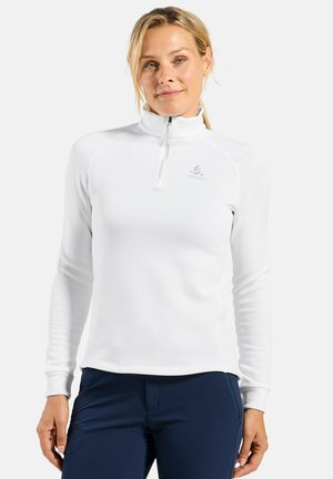 Vrouw draagt een witte lange mouwen zip-up top met een hoge kraag en marineblauwe broek, staand tegen een effen achtergrond.