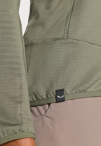 Top de manga larga verde oliva con un acabado texturizado y una etiqueta negra con un logo en el dobladillo, combinado con una parte inferior de tela beige claro.