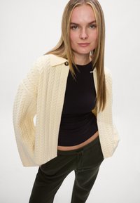 Kremowy kardigan w splot warkoczowy z szerokim kołnierzem i detalem w postaci guzików, zestawiony z dopasowanym czarnym topem i zielonymi spodniami ze sznurkiem.