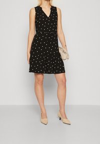 Robe noire sans manches à pois blancs, encolure en V, détail froncé aux épaules, ourlet à étages, associée à une petite pochette beige et des talons beiges.