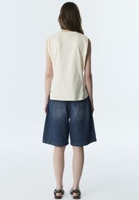 Mujer de espaldas, vistiendo una camiseta sin mangas beige, pantalones cortos sueltos de mezclilla oscura y sandalias negras sobre un fondo claro y liso.