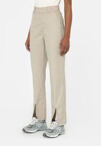 Osoba v béžových kalhotách s vysokým pasem a předními rozparky na lemu, s šedými teniskami New Balance, stojící na bílém pozadí.