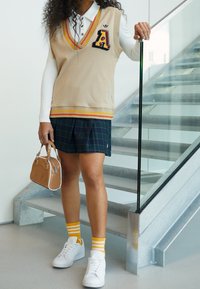 Beige stickad väst med broderad bokstav A, kombinerad med en marinblå rutig plisserad kjol. Vita sneakers och randiga gula strumpor fulländar looken.