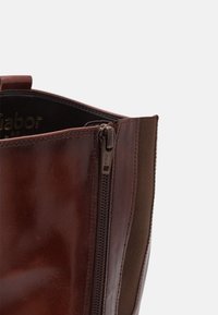 Gros plan sur une botte en cuir marron avec une fermeture éclair YKK visible et un panneau latéral élastique sur un fond uni clair.