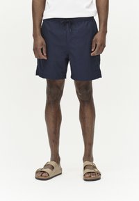 Dunkelblaue Badehose mit elastischem Bund und Kordelzug. Getragen mit beigen Schlupfsandalen mit einem doppelten Riemendesign.