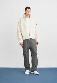 Crèmekleurig fleecejack met hoge kraag, grijze cargobroek met zakken en witte sneakers op een blauw tapijt tegen een effen witte achtergrond.