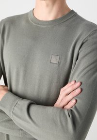 Henkilö, joka on pukeutunut vaaleanharmaaseen pitkähihaseen neulepaitaan, jossa on pieni neliön muotoinen "BOSS"-logo rinnassa, käsivarret ristissä edessä.