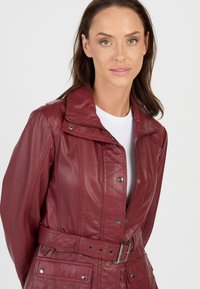 Kvind med lys hud og brunt hår iført en bælteholdt maroon læderjakke over en hvid top, vender fremad mod en ensfarvet baggrund.