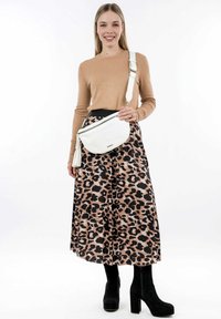 Bílá crossbody taška s zakřiveným designem a detailem v podobě třásní. Modelka má na sobě béžový svetr a sukni s leopardím potiskem, doplněnou černými botami na podpatku.